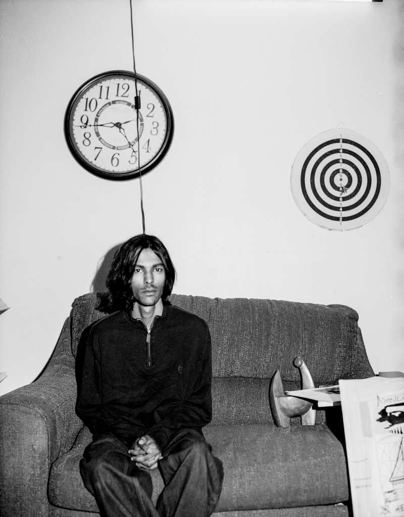 Seated on a couch beneath a wall clock and a dartboard